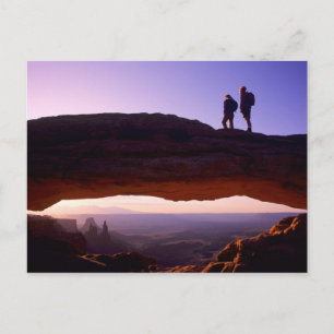 A couple watches sunrise from atop Mesa Arch in Postcard