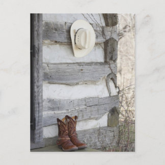 Cowboy boots and hat outside of log cabin postcard