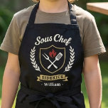 Custom Name Sous Chef Sidekick Child Apron<br><div class="desc">Make kitchen time extra fun with this personalized Sous Chef Sidekick kids apron. Featuring a stylish crest design with cooking utensils, flame detail, and custom name personalization, this apron is perfect for little helpers who love baking, cooking, and BBQ time with family. A great gift for birthdays, holidays, Father’s Day,...</div>
