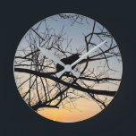 Golden Sunset Silhouette: Bird on Tree Branches Round Clock<br><div class="desc">Capture the serene beauty of twilight with this stunning silhouette photograph. A lone bird rests among the intricate web of bare tree branches, set against a vibrant golden and blue sunset sky. The high contrast and natural geometry create a peaceful, artistic atmosphere that complements any modern decor. This design is...</div>