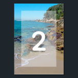 Rocky Beach Wedding Table Number<br><div class="desc">This beach design features a rocky shoreline. The picturesque setting combines greenery, blue ocean, rocky coastline and golden sand. The wording is elegantly framed with a simple white opaque border background. This is the perfect beach destination wedding design ideal for a summer or seaside wedding. Elegant and romantic beach wedding...</div>