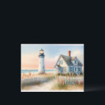 Seaside Lighthouse & Cottage Watercolor Scene Postcard<br><div class="desc">This charming coastal cottage watercolor captures the serene beauty of seaside living, featuring a classic white lighthouse standing tall beside a weathered beach house with blue shutters and a red brick chimney. Gentle waves roll towards the shore beneath a pastel sky, while a sailboat drifts peacefully in the distance. The...</div>