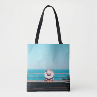 Woman wearing white straw hat sitting on bench loo tote bag