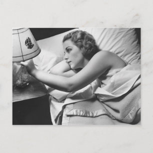 Young woman lying on bed turning off lamp on postcard